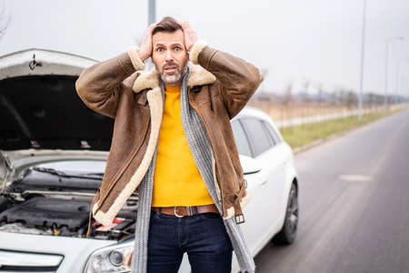 A man by the side of the road is standing in front of a broken car, waiting for the insurance...