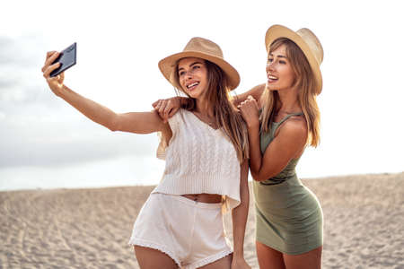 Two happy blonde caucasian sisters having fun on the beach during summer, taking selfie with mobile phone, smiling and looking to the smartphone camera. Beautiful girls wearing hats. Friendship. Real people emotions.の写真素材