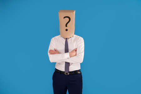 Businessman with brown paper bag on head with question mark, standing on blue studio background, a lot of copy space.の写真素材
