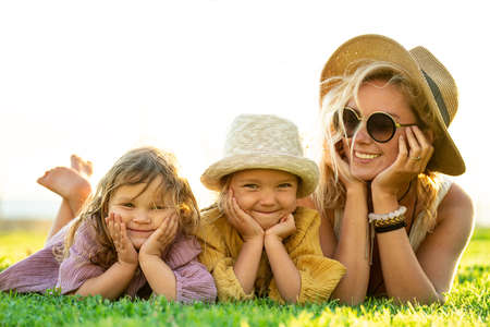 Beautiful photo of cute happy little sisters playing with her mother outside. Woman and her smiling daughters lying on the green grass in the park. Happy family. Happy Mother's Day. Motherhood, childhood. Summer vibes.の写真素材
