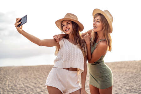 Two happy blonde caucasian sisters having fun on the beach during summer, taking selfie with mobile phone, smiling and looking to the smartphone camera. Beautiful girls wearing hats. Friendship. Real people emotions.の写真素材