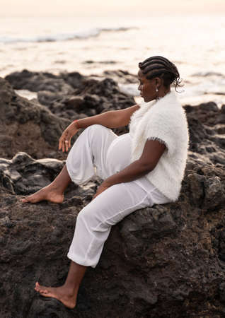 Beautiful pregnant woman in white clothes posing on the rocky coast, sitting and relaxing during sunset. Future motherhood concept. Mom to be. Outdoor photo.の写真素材