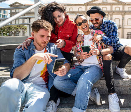 Happy group of multiracial students, friends using smartphone outdoors, watching social media and laughing, having fun together.の写真素材