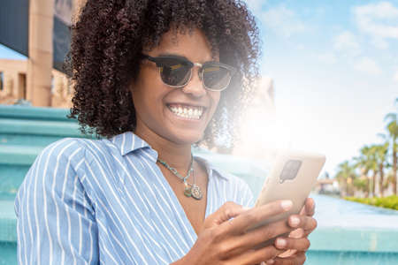 Portrait of happy young woman with afro hairstyle using mobile phone, having fun watching social media or talking online with friends. Girl wearing fashionable sunglasses. Outdoor photo.の写真素材