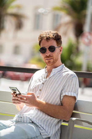 Casual handsome man using mobile phone outdoors, watching social media sitting on the bench in the city, relaxing.の写真素材