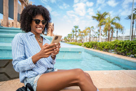 Happy fashionable woman using mobile phone at the city street, scrolling social media and laughing. Girl with afro hairstyle. Tourist. Travel concept.の写真素材