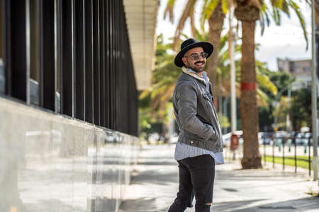 Fashionable, handsome man walking on the city street wearing black hat, sunglasses and jeans jacket, smiling to the camera.の写真素材