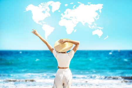 Conceptual photo of a women wearing straw hat, enjoying sunny time at the beach. Back view. World map shaped clouds in the blue sky. Travel. Tourism. A lot of copy space.の写真素材