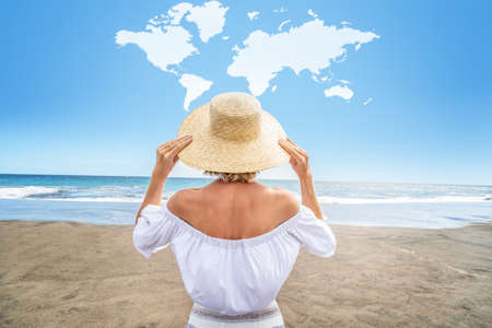 Conceptual photo of a women with straw hat enjoying sunny time at the beach. Back view. World map shaped clouds in the blue sky. Travel. Tourism. A lot of copy space.の写真素材