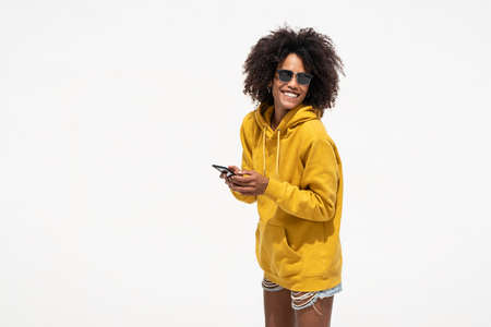 Smiling happy woman with afro hairstyle using smart phone, texting in social media. A lot of copy space. White wall.の写真素材