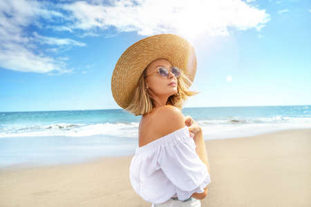 Portrait of blonde caucasian girl in summer straw hat and fashionable sunglasses walking, relaxing at the beach. Vacation vibe. Sunny day. Travel, tourism concept. A lot of copy space.の写真素材