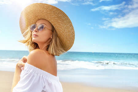 Portrait of blonde caucasian girl in summer straw hat and fashionable sunglasses walking, relaxing at the beach. Vacation vibe. Sunny day. Travel, tourism concept. A lot of copy space.の写真素材
