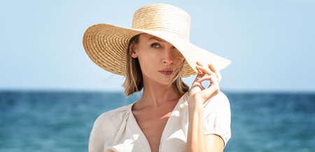 Beauty portrait of beautiful blonde caucasian girl in summer straw hat posing at the beach. Vacation vibe. Sunny day. Travel, tourism concept. A lot of copy space.の写真素材