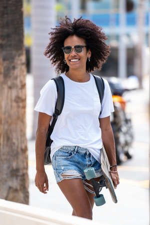 Young woman with afro hairstyle walking on the city street, carrying a skateboard, wearing fashionable sunglasses. Summer vibes. Hobby. Real people lifestyle.の写真素材
