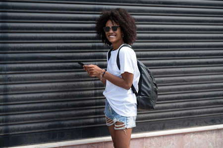 Happy attractive girl in the city street using mobile phone, checking social media and emails. Woman with afro hairstyle in stylish fashionable clothes. Real people emotions.の写真素材