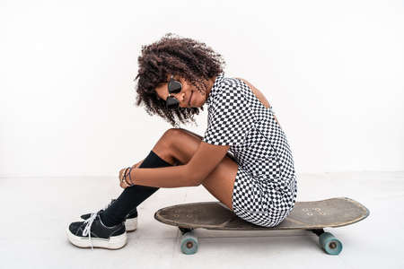 Photo of fashionable skater girl with afro hairstyle sitting on the skateboard, posing on the white...