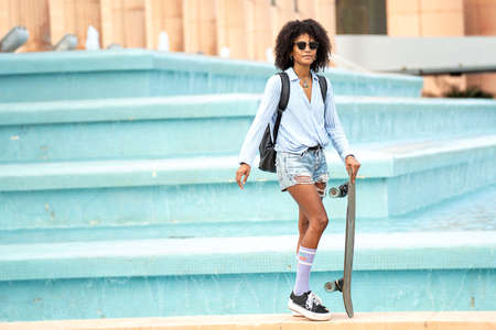 Young woman with afro hairstyle posing on the city street, carrying a skateboard, wearing...
