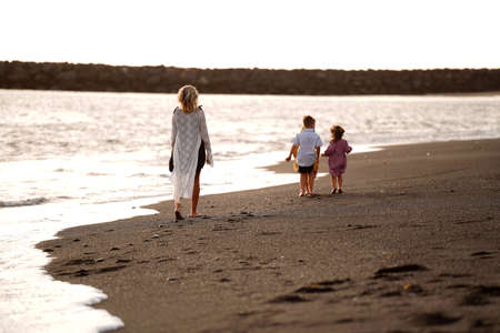 Joyful mother with three kids walking on the beach during sunset. Summer vacations with children. Back view. Concept of friendly family time on holidays. A lot of copy space.の写真素材