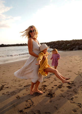 Joyful mother with two daughters playing on the beach, having fun during sunset, relaxing. Summer vacations with children. Concept of friendly family time on holidays. A lot of copy space.の写真素材