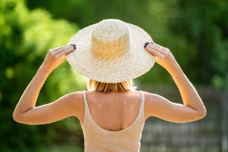 Woman in straw summer hat standing and looking at the green trees. Summer time. Back view. A lot of...