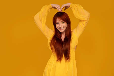 Gorgeous ginger woman in yellow elegant dress smiling to the camera, showing heart symbol. Female bright look. Colorful background and makeup. A lot of copy space.の写真素材