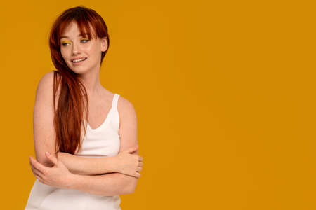 Cute natural redhead woman with delicate smile and yellow makeup posing on colorful studio background, looking away. Casual simple clothes. A lot of copy space.の写真素材