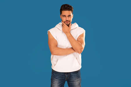 Fashionable young handsome man posing in white tank top with hood on blue studio background. Bearded guy looking at the camera. Copy space.の写真素材