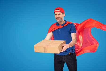 Happy handsome delivery man in red superhero cape and cap carrying cardboard box. Professional delivery service . Blue studio background. A lot of copy space.の写真素材