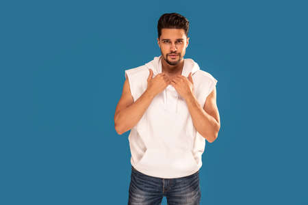 Fashionable young handsome man posing in white tank top with hood on blue studio background. Bearded guy looking at the camera. Copy space.の写真素材