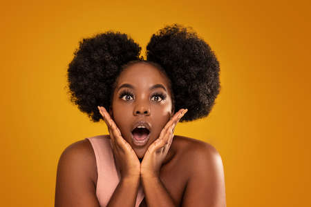 Portrait of funny young girl with afro hairstyle and long lashes making faces to the camera. Real people emotions, expression. Shocked woman on colorful yellow studio background. A lot of copy space for your text.の写真素材