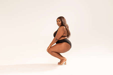 Attractive sexy dark skinned woman in black fashionable one swimsuit, looking at the camera. Full photo of superb plus size girl posing in studio on the gray background. Copy space. Concept of body acceptance, body positivity. Love yourself.の写真素材