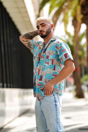 Photo of young man with beard and tattoos wearing fashionable colorful shirt and jeans, posing outdoor. Sensual guy.の写真素材
