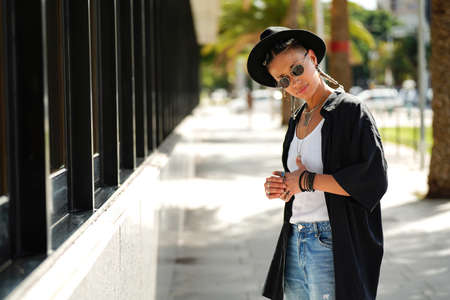 Fashionable hipster girl wearing black hat, shirt, jeans, sunglasses and jewelry posing on the city walk, looking at the camera. Sunny day.の写真素材