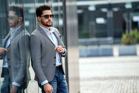 Outdoor photo of handsome elegant man wearing fashionable clothes and sunglasses, posing on the wall with reflections. A lot of copy space. City constantly. A small model.の写真素材
