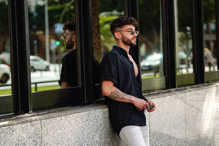 Street style. Fashionable handsome man in black shirt and sunglasses holding mobile phone in hands, waiting for somebody in the city.の写真素材