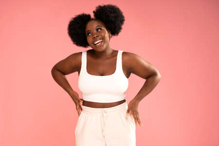 Attractive female model with dark skin and afro hairstyle posing on pink pastel studio background, smiling and showing her belly. Diet concept. Body positive, conscious. Real people emotions.の写真素材