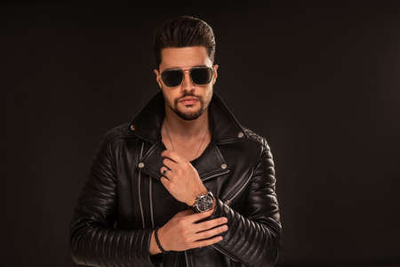 Fashion photo of handsome man wearing black leather jacket, sunglasses, hand watch, posing on dark studio background. Copy space.の写真素材
