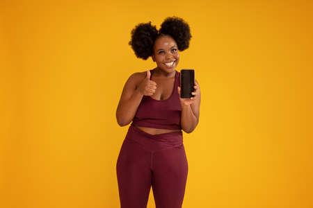 Smiling, happy, young girl with afro hairstyle posing on yellow studio background in sporty clothes, holding mobile phone in one hand showing new fitness app. Okay sign. Sale concept. Copy space.の写真素材