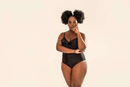 Seductive african woman in black fashionable swimsuit, posing looking at camera on gray studio background. Natural female beauty concept. Afro hairstyle. Plus size lady. Body positive.の写真素材