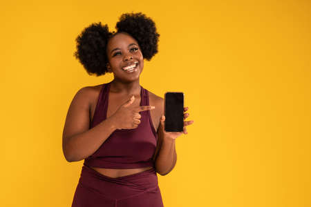 Smiling, happy, young girl with afro hairstyle posing on yellow studio background in sporty clothes, holding mobile phone in one hand showing new fitness app. Sale concept. Copy space.の写真素材