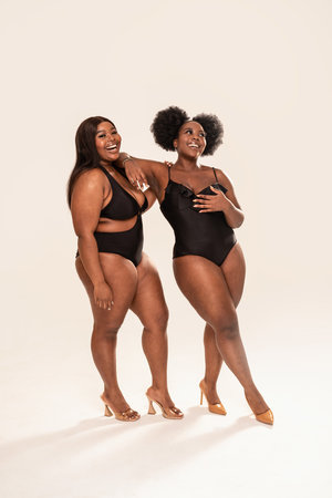 Two smiling, beautiful female models in black fashionable swimsuit posing together in studio, having fun together. Body positive concept. Plus size women. Copy space.の写真素材