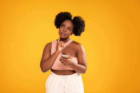 Young afro woman holding mobile phone and thinking about buying something online. Sale concept. Studio shot. Yellow background.の写真素材