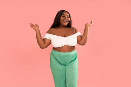 Joyful Plus Size african Woman laughing, posing on a pastel pink Studio Background. Real people emotions, toothy smile. Fashionable young girl in white top and green pants. Body positive. A lot of copy space.の写真素材