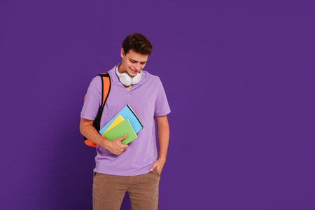 Young happy man student in casual clothes posing with backpack and notebooks, isolated on studio violet background. Education concept. Copy space.の写真素材