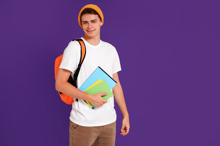 Young happy man student in casual clothes and cap, with backpack and notebooks, isolated on studio violet background. Education concept. Copy space.の写真素材