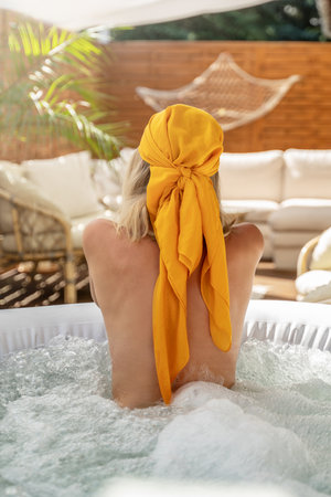 Back view of a woman relaxing in a bathtub. Girl wearing fashionable head scarf. Real summer lifestyle, wellness spa concept. Boho terrace at home. Sunny day.の写真素材