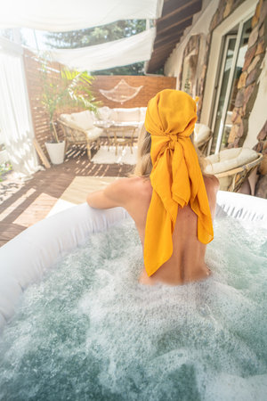 Back view of a woman relaxing in a bathtub. Girl wearing fashionable head scarf. Real summer lifestyle, wellness spa concept. Boho terrace at home. Sunny day.の写真素材