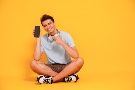 Happy smiling young man in casual clothes showing empty mobile phone screen, isolated on yellow studio background. People lifestyle concept. Copy space.の写真素材