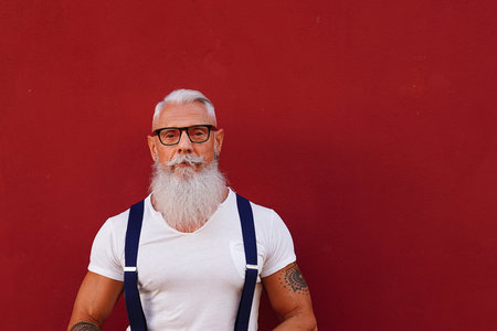 Portrait of casual, confident senior with long white beard and mustache and tattoos. Man wearing shirt and suspenders, looking at camera, isolated on red background. A lot of copy space.の写真素材