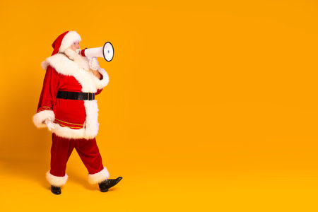 Christmas is coming! Real Santa Claus shouting using megaphone over yellow studio background. Xmas sale, discount concept. Full length photo.の写真素材